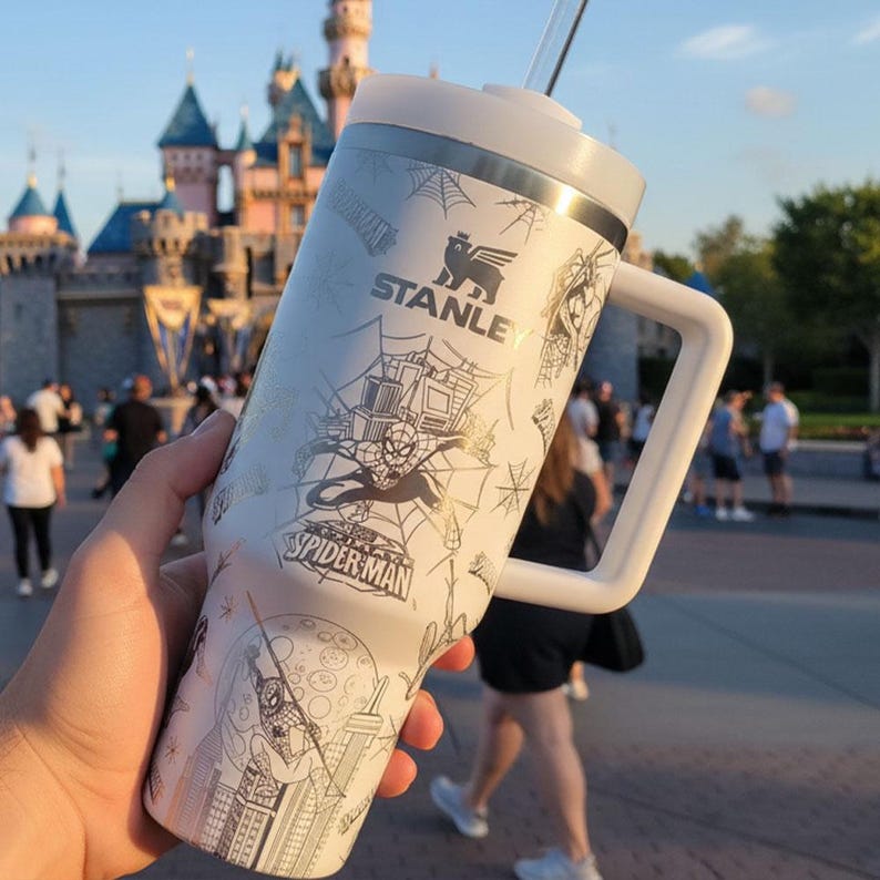 Pu&ograve; includere: Un bicchiere Stanley bianco con manico, con grafica di Spider-Man e il nome del personaggio. Il design include ragnatele e paesaggi urbani. Il bicchiere ha un coperchio bianco e una cannuccia trasparente.