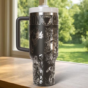 May include: Black stainless steel travel mug with a white lid and handle. The mug features a detailed silver graphic of Iron Man and the words "IRON MAN". The mug is designed to keep beverages hot or cold.