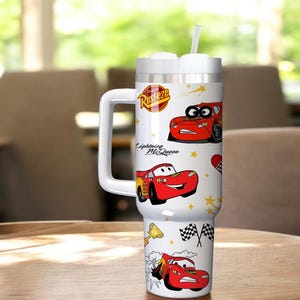 May include: A white tumbler with a handle, featuring illustrations of the character Lightning McQueen from the movie Cars. The tumbler has a white lid with a straw and the text "Rusteze" and "Lightning McQueen".