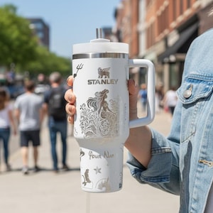 May include: A white Stanley tumbler with a handle, lid, and straw. The tumbler features a mermaid design, the Stanley logo, and the words "Your Name" in a script font. The background is a blurred street scene.