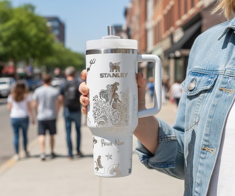 May include: A white Stanley tumbler with a handle, lid, and straw. The tumbler features a mermaid design, the Stanley logo, and the words "Your Name" in a script font. The background is a blurred street scene.