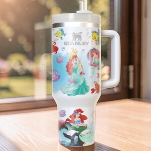 Puede incluir: Vaso Stanley blanco con ilustraciones de Ariel y otros personajes de La Sirenita. El vaso tiene un asa blanca, una tapa plateada y una pajita. El dise&ntilde;o incluye a Ariel, Flounder, Sebasti&aacute;n y otras criaturas marinas.