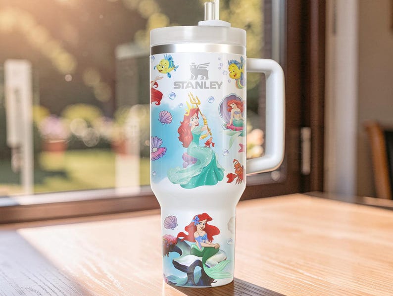 Puede incluir: Vaso Stanley blanco con ilustraciones de Ariel y otros personajes de La Sirenita. El vaso tiene un asa blanca, una tapa plateada y una pajita. El dise&ntilde;o incluye a Ariel, Flounder, Sebasti&aacute;n y otras criaturas marinas.