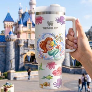 Puede incluir: Taza de viaje Stanley blanca con una colorida ilustraci&oacute;n de Ariel de La Sirenita, con Flounder y Sebasti&aacute;n. La taza tiene un asa y tapa blancas y est&aacute; decorada con criaturas marinas. El logotipo de Stanley es visible.