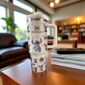 May include: White Stanley travel mug featuring a repeating pattern of the character Stitch from Lilo & Stitch. The mug has a white handle and lid, with the Stanley logo and Stitch graphics in blue and purple. The mug is on a wooden table.