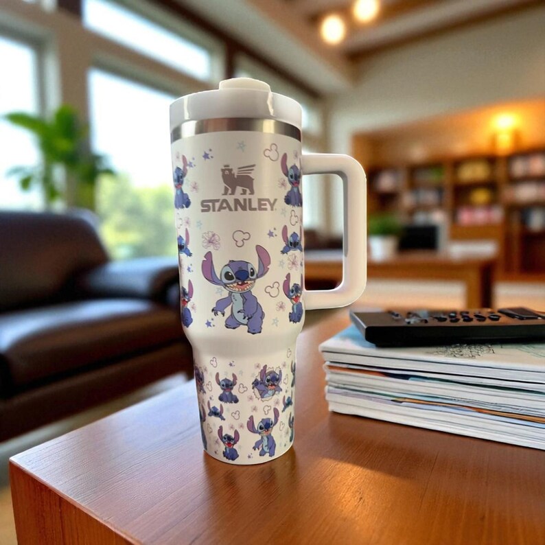 May include: White Stanley travel mug featuring a repeating pattern of the character Stitch from Lilo & Stitch. The mug has a white handle and lid, with the Stanley logo and Stitch graphics in blue and purple. The mug is on a wooden table.