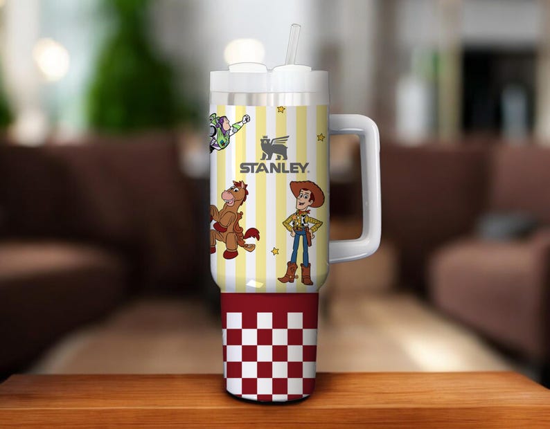 May include: A white and yellow Stanley tumbler with a handle, featuring cartoon characters and the Stanley logo. The bottom section has a red and white checkered pattern. Includes a white lid and straw.