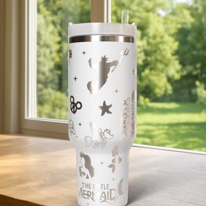 May include: White insulated tumbler with a silver design featuring characters and icons from "The Little Mermaid." The tumbler has a white lid and a clear straw. The design includes a silhouette of King Triton.