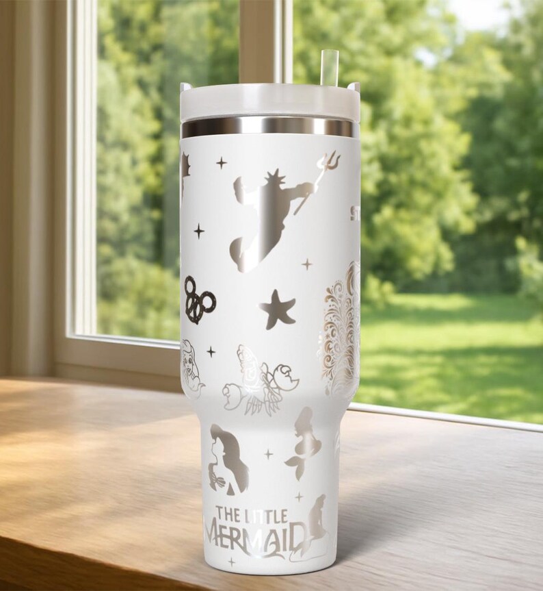 May include: White insulated tumbler with a silver design featuring characters and icons from "The Little Mermaid." The tumbler has a white lid and a clear straw. The design includes a silhouette of King Triton.