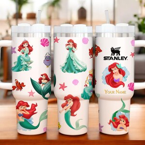 Puede incluir: Vaso t&eacute;rmico blanco con ilustraciones de Ariel de La Sirenita, con asa y tapa blancas. El vaso incluye una pajita y el logotipo de Stanley. El dise&ntilde;o incorpora conchas marinas, estrellas de mar y el texto "Your Name".