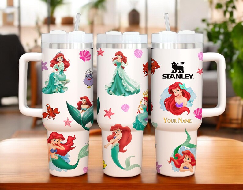 Puede incluir: Vaso t&eacute;rmico blanco con ilustraciones de Ariel de La Sirenita, con asa y tapa blancas. El vaso incluye una pajita y el logotipo de Stanley. El dise&ntilde;o incorpora conchas marinas, estrellas de mar y el texto "Your Name".