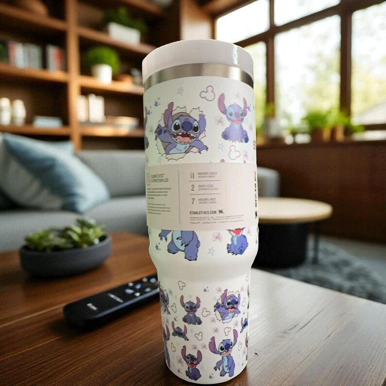 May include: A white insulated tumbler featuring a repeating pattern of the character Stitch from Lilo & Stitch. The tumbler has a white lid and a black and white label with text. The tumbler is on a wooden table.