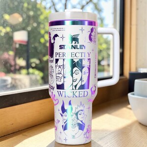Puede incluir: Vaso Stanley blanco con asa y tapa blancas, con un dise&ntilde;o de villanos de Disney. El vaso tiene las palabras "Stanley", "Perfectly Wicked" y otro texto. El dise&ntilde;o incluye detalles en morado e iridiscente.