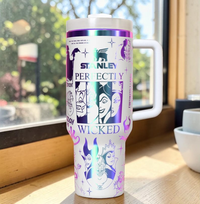 Puede incluir: Vaso Stanley blanco con asa y tapa blancas, con un dise&ntilde;o de villanos de Disney. El vaso tiene las palabras "Stanley", "Perfectly Wicked" y otro texto. El dise&ntilde;o incluye detalles en morado e iridiscente.