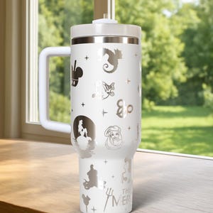 May include: White insulated tumbler with a handle, lid, and straw. The tumbler is decorated with silver-colored illustrations of characters and symbols, including a seahorse, a mermaid, and the text "THE MER".
