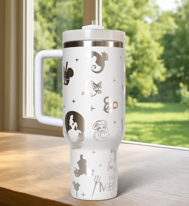 May include: White insulated tumbler with a handle, lid, and straw. The tumbler is decorated with silver-colored illustrations of characters and symbols, including a seahorse, a mermaid, and the text "THE MER".