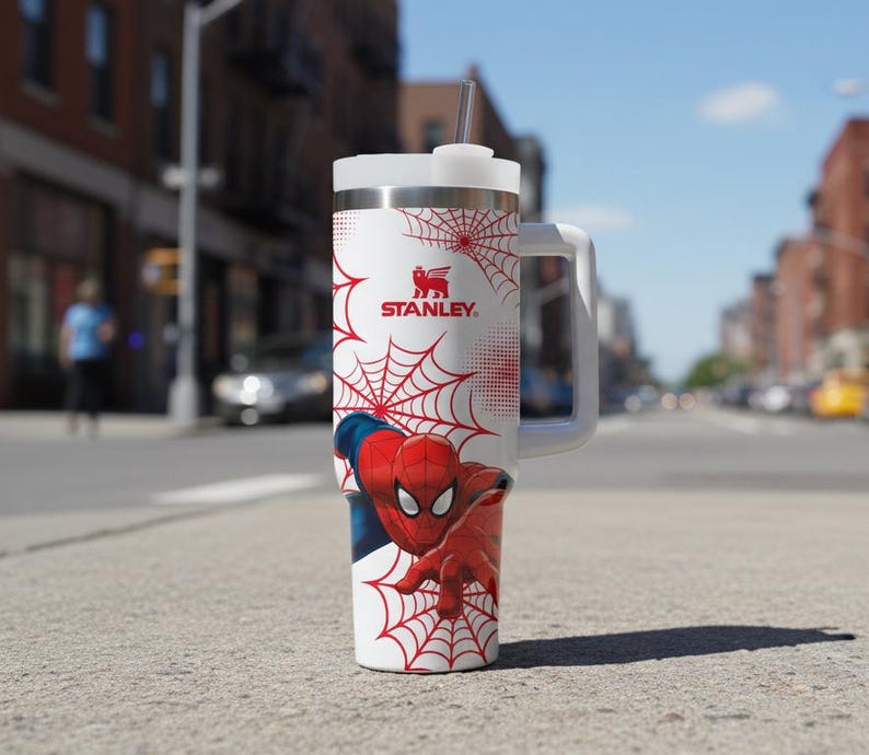 May include: A white and stainless steel Stanley tumbler featuring Spider-Man graphics. The tumbler has a red spiderweb design, the Spider-Man character, and the Stanley logo. It includes a white handle, lid, and a straw.