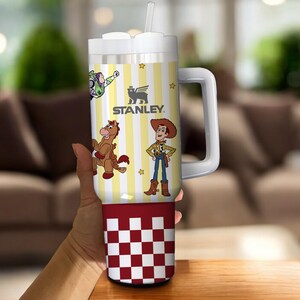 May include: A white and yellow Stanley tumbler with a handle, featuring cartoon characters and the Stanley logo. The bottom section has a red and white checkered pattern. Includes a straw and a white lid.