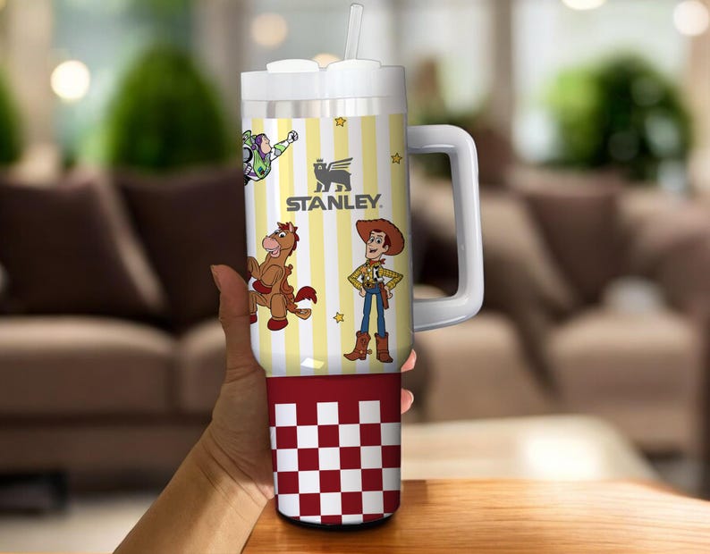 May include: A white and yellow Stanley tumbler with a handle, featuring cartoon characters and the Stanley logo. The bottom section has a red and white checkered pattern. Includes a straw and a white lid.