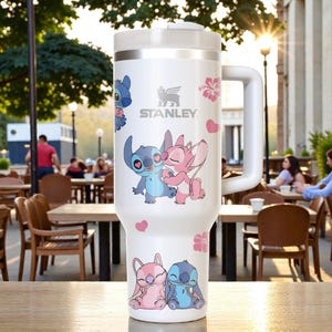 Puede incluir: Vaso Stanley blanco con asa, con los personajes de dibujos animados Stitch y Angel, junto con corazones y flores rosas. El logotipo de Stanley es visible. El vaso est&aacute; sobre una superficie de madera.