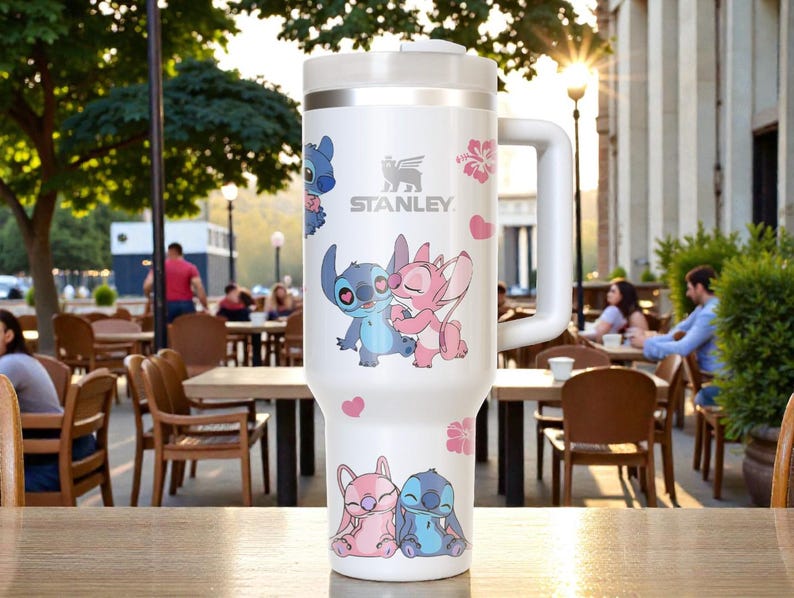 Puede incluir: Vaso Stanley blanco con asa, con los personajes de dibujos animados Stitch y Angel, junto con corazones y flores rosas. El logotipo de Stanley es visible. El vaso est&aacute; sobre una superficie de madera.