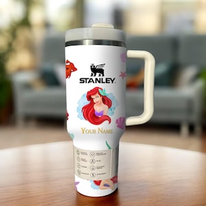 Puede incluir: Vaso Stanley blanco con tapa y asa grises, con una ilustraci&oacute;n de dibujos animados de Ariel de La Sirenita, un cangrejo rojo y el texto "Your Name". El vaso tiene un logotipo negro de Stanley.