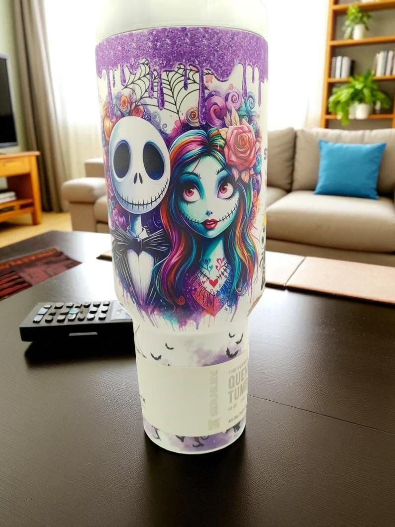 May include: A white tumbler featuring Jack Skellington and Sally from The Nightmare Before Christmas. The design includes a purple glitter top, dripping accents, and a floral arrangement. The tumbler has a white band with text.