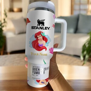 Puede incluir: Vaso Stanley Quencher H2.0 Flowstate blanco con asa, con una ilustraci&oacute;n de dibujos animados de Ariel de La Sirenita. El vaso tiene el logotipo de Stanley y el texto "Your Name".