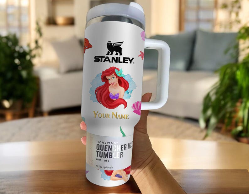 Puede incluir: Vaso Stanley Quencher H2.0 Flowstate blanco con asa, con una ilustraci&oacute;n de dibujos animados de Ariel de La Sirenita. El vaso tiene el logotipo de Stanley y el texto "Your Name".