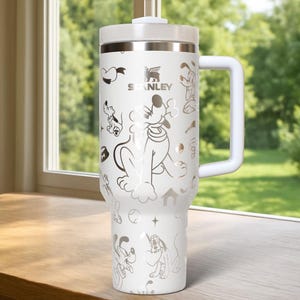 May include: White insulated tumbler with a handle and lid. The tumbler features a silver design of Pluto, hearts, bones, and other dog-themed graphics. The Stanley logo is visible. The tumbler is sitting on a wooden surface.