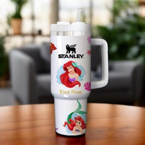 Puede incluir: Vaso Stanley blanco con asa, pajita y tapa. Presenta una imagen de dibujos animados de Ariel de La Sirenita, con el texto "Your Name" debajo. El logotipo de Stanley tambi&eacute;n es visible.
