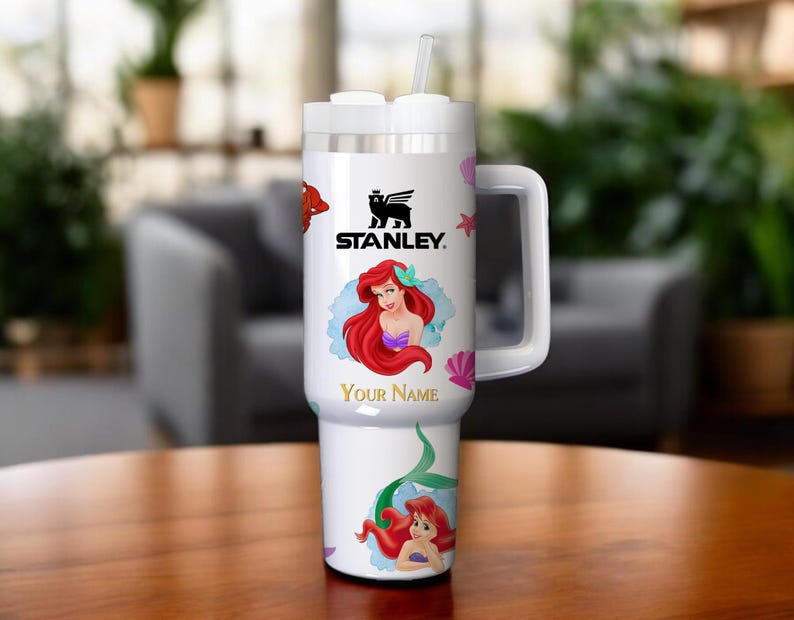 Puede incluir: Vaso Stanley blanco con asa, pajita y tapa. Presenta una imagen de dibujos animados de Ariel de La Sirenita, con el texto "Your Name" debajo. El logotipo de Stanley tambi&eacute;n es visible.