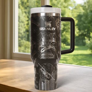 Pu&ograve; includere: Tazza da viaggio Stanley nera con coperchio bianco e manico nero. La tazza presenta un design di Spider-Man con il testo "STANLEY" e "SPIDER-MAN" in argento. Il design include ragnatele, paesaggi urbani e illustrazioni di Spider-Man.