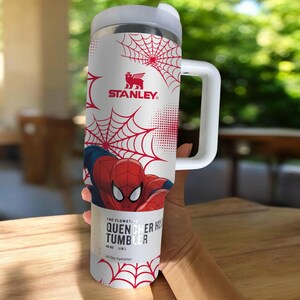 May include: A white Stanley Quencher H2.0 Flowstate tumbler featuring Spider-Man graphics and red spiderwebs. The tumbler has a handle and a white lid. The text on the tumbler reads "THE FLOWSTATE QUENCHER H2.0 TUMBLER" and "40 OZ | 1.18 L".