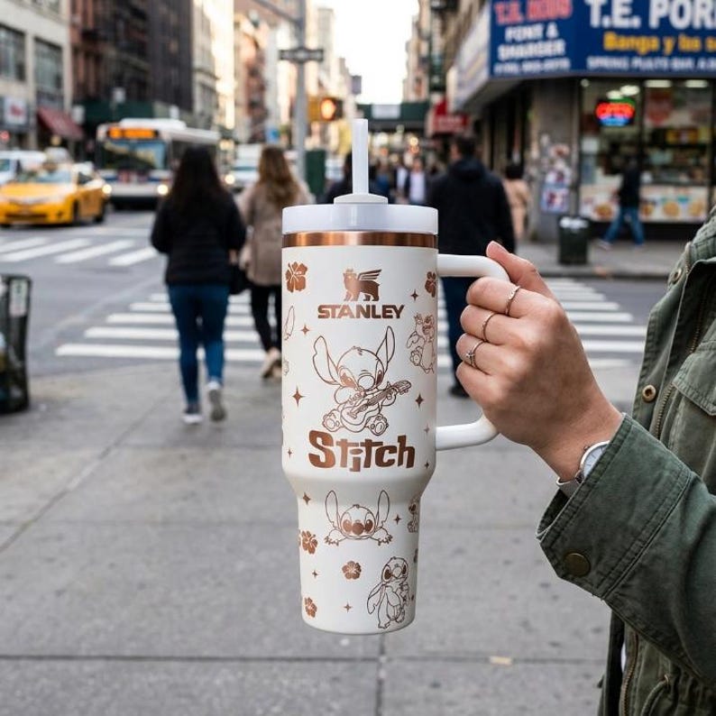 May include: A white Stanley tumbler with a copper-colored lid, handle, and Stitch graphics. The tumbler features the word "Stitch" and a graphic of Stitch playing a guitar. The background shows a city street.