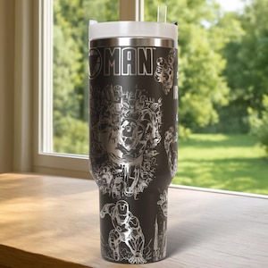 May include: A black stainless steel travel mug with a silver Iron Man design. The mug has a white lid and a clear straw. The words "IRON MAN" are visible, along with illustrations of the character in action. The mug is suitable for hot or cold drinks.