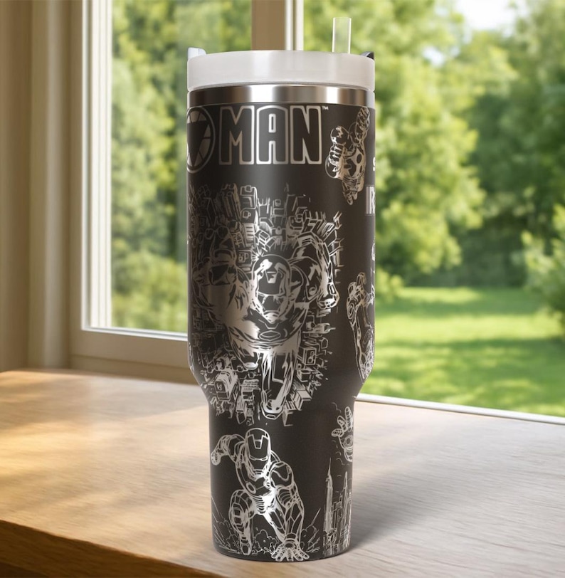 May include: A black stainless steel travel mug with a silver Iron Man design. The mug has a white lid and a clear straw. The words "IRON MAN" are visible, along with illustrations of the character in action. The mug is suitable for hot or cold drinks.