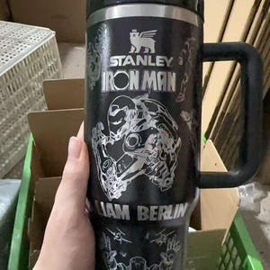 May include: Black Stanley Iron Man travel mug with a black handle and lid. The mug features a silver Iron Man graphic and the text "STANLEY IRON MAN" and "LIAM BERLIN".