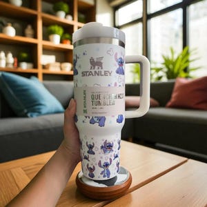 May include: A white Stanley Quencher H2.0 tumbler with a handle, featuring a Stitch character design. The tumbler has a white lid and handle, and the body is adorned with Stitch illustrations and the Stanley logo. It sits on a wooden coaster.