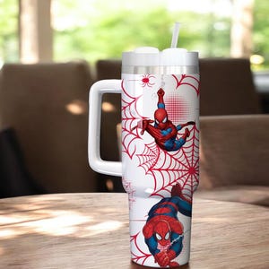 May include: A white insulated tumbler featuring Spider-Man graphics. The design includes red spiderwebs and illustrations of Spider-Man in action. The tumbler has a white handle and a straw.