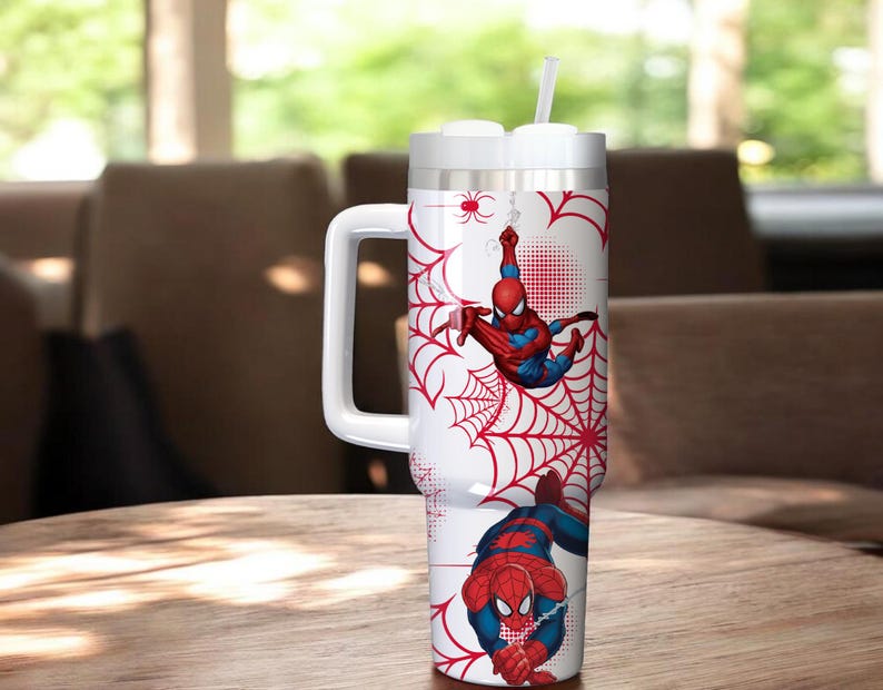 May include: A white insulated tumbler featuring Spider-Man graphics. The design includes red spiderwebs and illustrations of Spider-Man in action. The tumbler has a white handle and a straw.