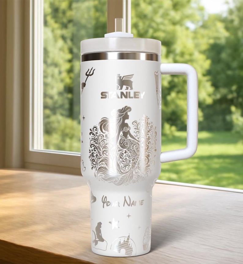May include: White Stanley tumbler with a handle, lid, and straw. The tumbler features a silver mermaid design, the Stanley logo, and the words "Your Name". Additional silver illustrations include a trident, castle, and stars.