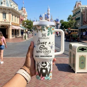 以下が含まれることがあります： 白いハンドル付きスタンレータンブラー。花柄と蝶のデザインが施されています。「Just a Girl Who Loves Jack Skellington」の文字とジャック・スケリントンのグラフィックがあります。白い蓋とストローが含まれています。