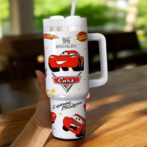 May include: White Stanley tumbler featuring Lightning McQueen from the Cars movie. The tumbler has a large white handle and a white lid with a straw. The Cars logo and the Stanley logo are also visible on the tumbler.