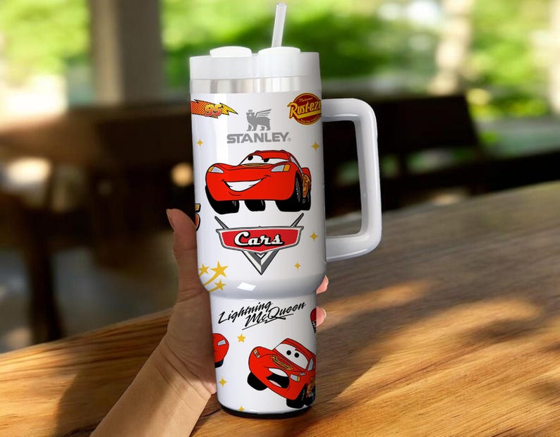 May include: White Stanley tumbler featuring Lightning McQueen from the Cars movie. The tumbler has a large white handle and a white lid with a straw. The Cars logo and the Stanley logo are also visible on the tumbler.
