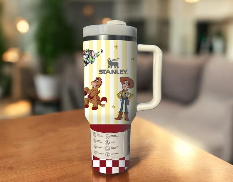May include: A Stanley travel mug with a white handle and lid. The mug features a yellow and white striped pattern with cartoon characters and the Stanley logo. The bottom of the mug has a red and white checkered pattern.