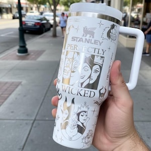 Puede incluir: Taza de viaje Stanley blanca con tapa y asa plateadas. La taza presenta las palabras "Stanley Perfectly Wicked" e ilustraciones de villanos de Disney. La taza est&aacute; sujeta por una persona.