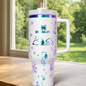 May include: White Stanley tumbler with a handle, featuring Snoopy and other Peanuts characters in iridescent colors. The tumbler has a white lid and a straw. The Stanley logo is visible. The tumbler is designed for cold beverages.