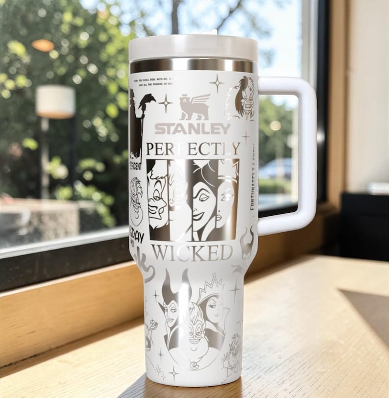 Puede incluir: Un vaso Stanley blanco con asa y tapa blancas. El vaso presenta ilustraciones de villanos de Disney y las palabras "Stanley Perfectly Wicked". El vaso est&aacute; dise&ntilde;ado para mantener las bebidas calientes o fr&iacute;as.