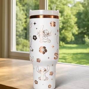 May include: White tumbler with a rose gold band and lid, featuring a repeating pattern of the character Stitch, flowers, and stars. The tumbler has a straw and the letter "S" on the side. The tumbler is sitting on a wooden surface.
